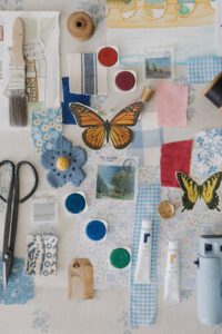 A close up of a blue-toned mood board with paints, brushes, fabric and other craft items.