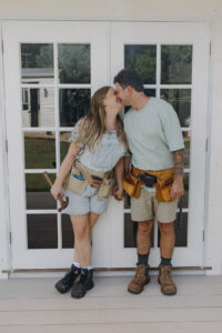A woman and man dressed in work clothes with tool belts, kissing.