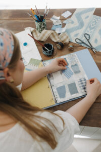 High angle over the shoulder of a woman placing material swatches in a folder with paints and other craft items in the shot.