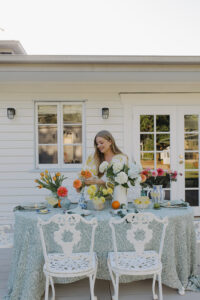 A woman smiling, styling flowers on an outdoor dining table with more fresh flowers, a blue floral tablecloth and white vintage chairs.