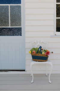 A cane basket filled with fresh flowers, sitting on a vintage white chair beside a blue door.