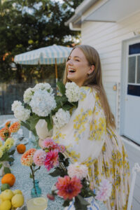 A woman smiling, holding white and purple flowers beside a styled outdoor dining table with more fresh flowers.