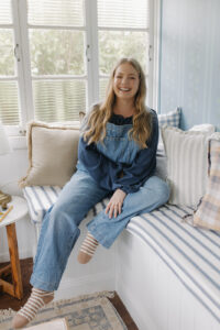 A woman in overalls, sitting on a blue and white striped window seat.