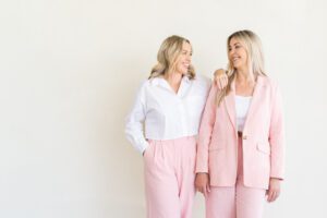 Nurse Jodie and Nurse Pru stand next to each other against a beige wall, they are both wearing pink and smiling.