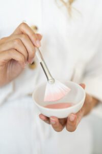 Hands holding a bowl of pink face mask and a brush dipped in.