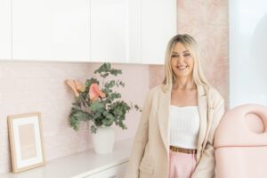 Nurse Jodie standing and smiling in a room featuring bespoke pink palm wallpaper, pink subway tiles, and a bouquet of fresh flowers.