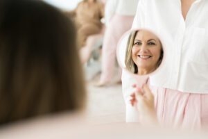 A beauty clinic client smiles at her reflection as she holds a pink handheld mirror.