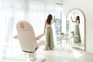 A nurse pulls back sheer curtains in a beauty clinic room to reveal waterfront views.
