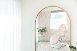 A pink beauty clinic chair, bespoke pink palm wallpaper, and a fresh bouquet of flowers are shown in the reflection of an arched brass mirror.
