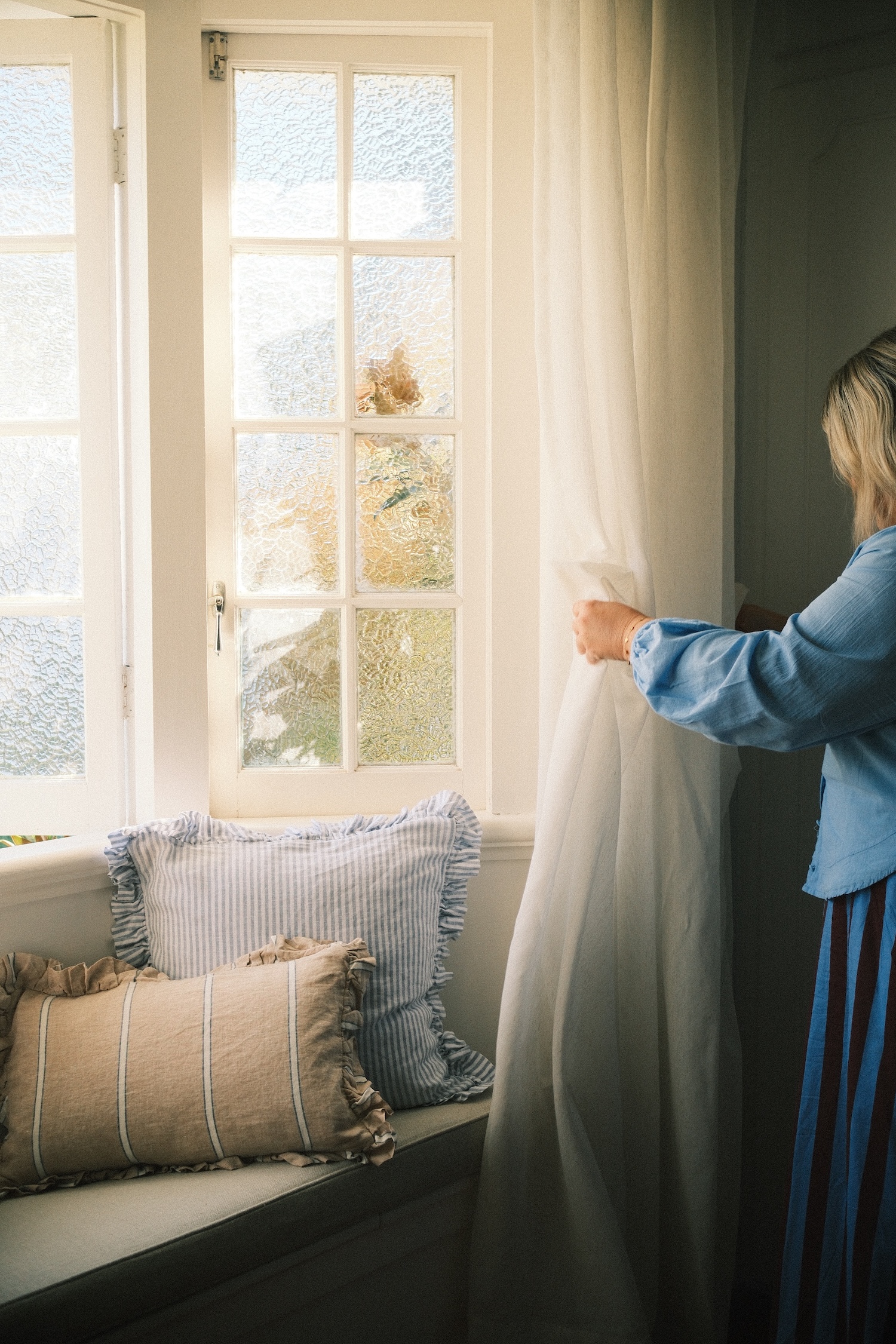 Blonde lady in a blue shirt, fixing the curtain of a window seat.