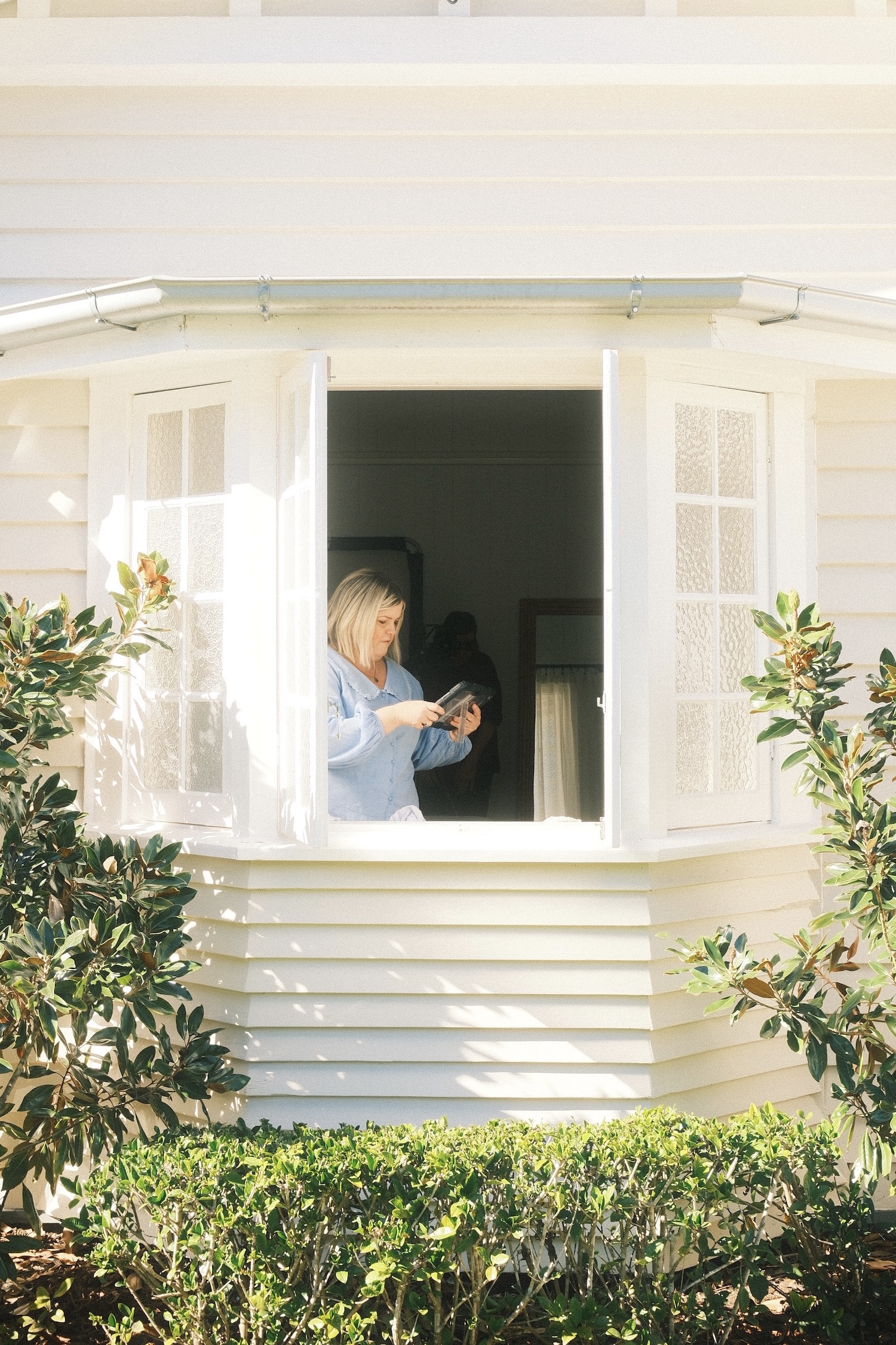 A lady looking at a tablet in the window of a white country style home.