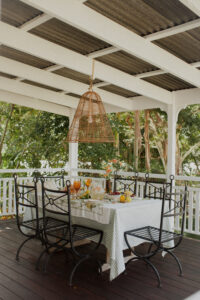 A festive spread covers an outdoor table on a renovated Queenslander veranda. There are fresh flowers, citrus fruits and bunches of grapes, and a pitcher of sparkling punch. Wrought iron dining chairs surround the table and a rattan pendant light hangs above it.