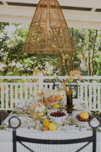 A festive spread covers an outdoor table on a renovated Queenslander veranda. There are fresh flowers, citrus fruits and bunches of grapes, and a pitcher of sparkling punch. Wrought iron dining chairs surround the table and a rattan pendant light hangs above it.