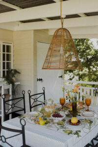 A festive spread covers an outdoor table on a renovated Queenslander veranda. There are fresh flowers, citrus fruits and bunches of grapes, and a pitcher of sparkling punch. Wrought iron dining chairs surround the table and a rattan pendant light hangs above it.