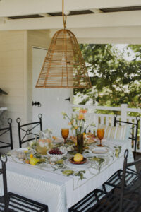 A festive spread covers an outdoor table on a renovated Queenslander veranda. There are fresh flowers, citrus fruits and bunches of grapes, and a pitcher of sparkling punch. Wrought iron dining chairs surround the table and a rattan pendant light hangs above it.