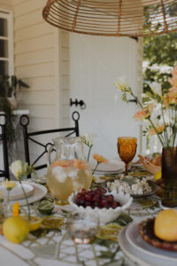 A festive spread covers an outdoor table on a renovated Queenslander veranda. There are fresh flowers, citrus fruits and bunches of grapes, and a pitcher of sparkling punch. Wrought iron dining chairs surround the table and a rattan pendant light hangs above it.
