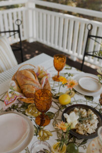 A festive spread covers an outdoor table on a renovated Queenslander veranda. There are fresh flowers, citrus fruits, potato salad, and a basket of sourdough sitting on a vintage tablecloth.