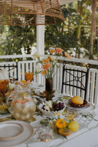 A festive spread covers an outdoor table on a renovated Queenslander veranda. There are fresh flowers, citrus fruits and bunches of grapes, and a pitcher of sparkling punch. Wrought iron dining chairs surround the table and a rattan pendant light hangs above it.