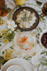 A close up of a festive spread. Looking down at a jug of lychee and elderflower sparkling punch, a bowl of potato salad, and fresh fruits and flowers sit upon a vintage tablecloth.