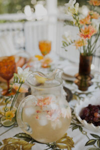 A close up of a festive spread. Looking at a jug of lychee and elderflower sparkling punch, fresh fruits and flowers sit upon a vintage tablecloth.