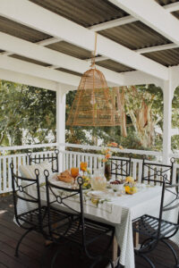 A festive spread covers an outdoor table on a renovated Queenslander veranda. There are fresh flowers, citrus fruits and bunches of grapes, and a pitcher of sparkling punch. Wrought iron dining chairs surround the table and a rattan pendant light hangs above it.
