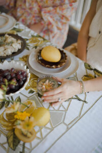 Looking down at a festive spread with fresh flowers and fruits on a vintage tablecloth, a hand holds a gold goblet of sparkling punch.