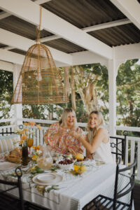 Stylist Hayley Jenkin and Sarah Bell happily cheers around a festive tabletop on a renovated Queenslander veranda. Wrought iron chairs surround the table and a rattan light pendant hangs above it.
