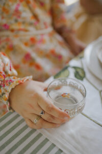 A close up of stylist Hayley Jenkin's hand grasping a vintage gold goblet filled with sparkling punch. She is wearing a floral La Boheme dress, a handmade sea glass heart ring, and a vintage gold constellation ring.