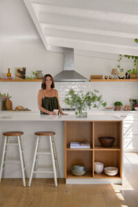 A woman standing at the bench of a white country style kitchen with stools and styled shelves.