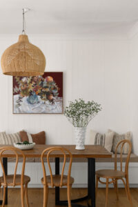 A wooden dining table and chairs with a vase and flowers on top, burgundy artwork on the wall, and a large rattan light shade.