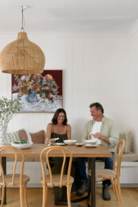 A woman and a man sitting at a dining table with burgundy artwork on the wall behind them and a large rattan light shade.