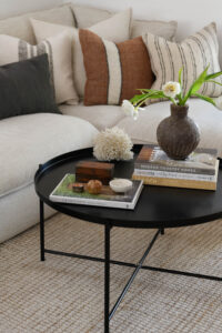 Close up shot of a black coffee table with books, vases and trinkets on top and a white lounge and cushions in the background.