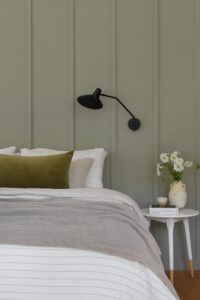 Sage green wall with a black reading lamp and thoughtfully styled bed and bedside table.