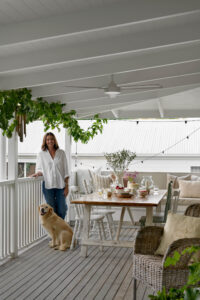 A lady in a white shirt with her dog, standing on the back verandah of a white, renovated country style Queensland home with eclectic chairs around a table with afternoon tea served upon it.