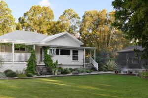 A wide exterior shot of a renovated white country style Queenslander home with green grass and a fire pit area to the right.