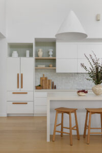 A modern kitchen with MCM House pendant, white cabinetry, and greenery in a vase on the bench.