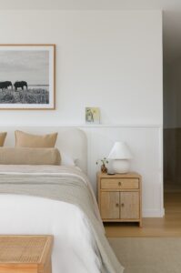 A modern primary suite with neutral ben linen and wooden bedside table with white lamp.