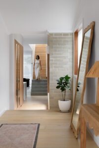 The entryway of a modern cottage, featuring grey brick and warm timber tones. The home owner is running down the stairs.