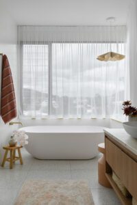 A free standing bathtub in a modern cottage ensuite, alongside a large window with sheer curtains.