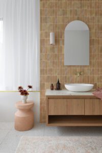 A vanity in a modern cottage bathroom, featuring terracotta coloured tiles, fresh flowers, and an arched mirror.