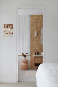 Looking through a doorway into a vanity in a modern cottage bathroom, featuring terracotta coloured tiles, fresh flowers, and an arched mirror.