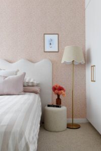 A little girl's room in a modern cottage, featuring baby pink floral wallpaper, a boucle ottoman, a scalloped detail floor lamp, and pink throw cushions.