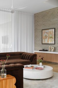 The living room of a modern cottage, featuring grey brick walls and floor to ceiling sheer curtains. The couch is a brown velvet Sarah Ellison sofa, there are handmade ceramics and fresh flowers on the coffee table.