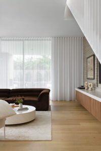 The living room of a modern cottage, featuring floor to ceiling sheer curtains and a brown velvet Sarah Ellison sofa. There are handmade ceramics and fresh flowers on the coffee table and sideboard.