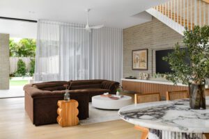 The living room of a modern cottage, featuring grey brick walls and floor to ceiling sheer curtains. The couch is a brown velvet Sarah Ellison sofa, there are handmade ceramics and fresh flowers on the coffee table, as well as an Italian marble dining table in the foreground.