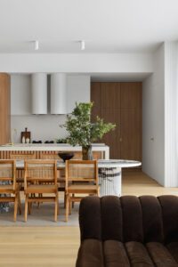 Looking through the dining area into the kitchen of a modern cottage. The dining table is Italian marble and has a big vase of tall fresh greenery in it, as well as timber rattan chairs surrounding it.