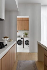 Looking through the kitchen of this modern cottage into the laundry. A washer and dryer sit side by side with timber cabinetry above.