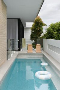 The pool area in the backyard of a modern cottage. There are two rattan pool chairs poolside and two blue and white stripy rings floating in the pool.