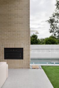 The outdoor area of a modern cottage. The exterior is brick, and there is a brass outdoor shower and built-in woodfire pizza oven.