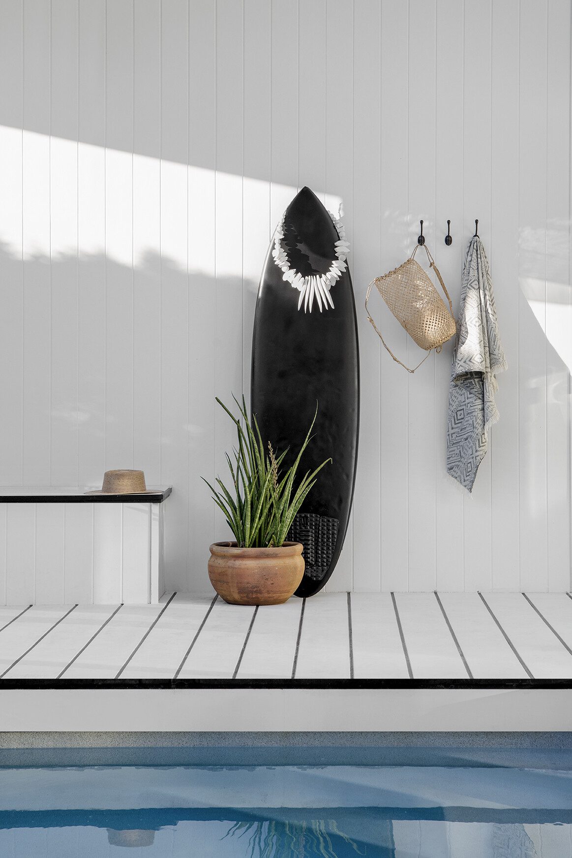 A black surfboard and towel lean up agains the wall near a pool.